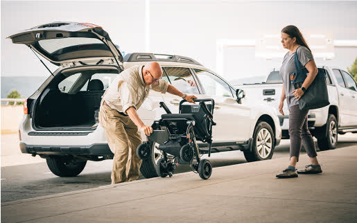 A man folding a Jazzy Carbon beside an open SUV while a companion stands nearby